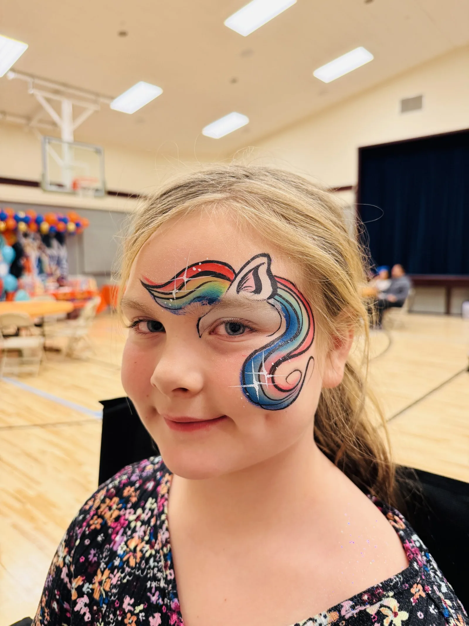 Child with rainbow unicorn cheek art at an indoor event.